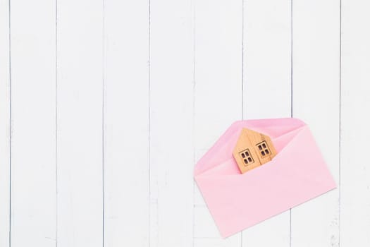 Flat lay of pink envelope with wooden house model on white table background for family, love, housing and property concept