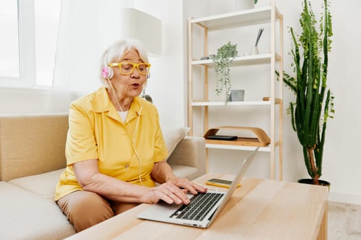 Happy elderly woman with a laptop typing with headphones sitting at home on the couch in a yellow shirt, bright modern interior, lifestyle online communication. High quality photo