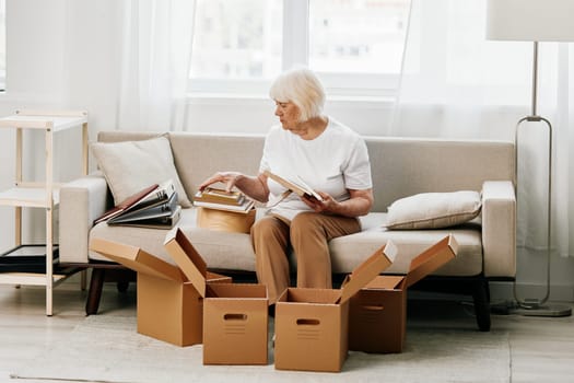elderly woman sits on a sofa at home with boxes. collecting things with memories albums with photos and photo frames moving to a new place cleaning things and a happy smile. Lifestyle retirement. High quality photo