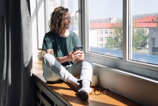 Digital Disconnect: Peaceful Young Woman Finds Serenity at Home, Gazing Out of Window, Balancing Technology and Relaxation in a Lockdown Era.