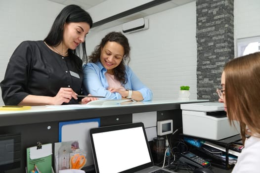 Experienced pleasant young female doctor dentist or beautician aesthetician in black uniform talking to her patient at reception desk, discussing the treatment that need to perform in medical clinic