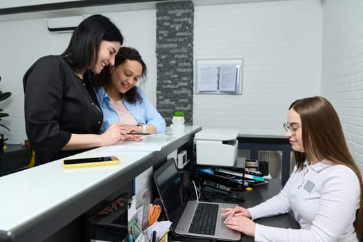 Female doctor and patient discussing medical treatment needs to perform, filling out and signing forms, standing at the reception desk, while the manager receptionist works on laptop in medical clinic