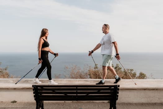 Happy Middle aged couple or friends practicing nordic walking in park near sea. Mature couple with trekking poles walking, practicing Nordic walking outdoors. Aging youthfully and sport concept.