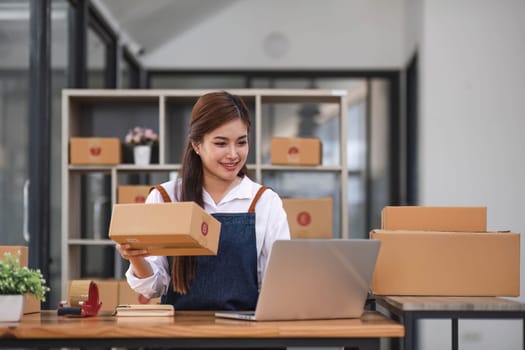 Startup happy Asian woman business owner works with a box at home office. prepare parcel delivery SME supply chain, procurement, package box to deliver to customers, Online SME business entrepreneurs ideas