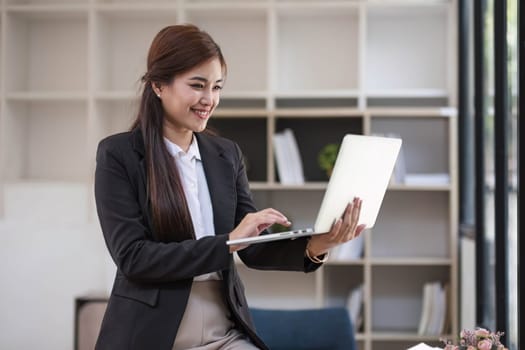 Portrait, Professional and confident millennial Asian businesswoman or female executive manager in formal suit and eyeglasses, standing, leaning on table, holding laptop and using laptop computer...