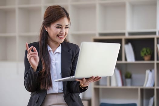 Portrait, Professional and confident millennial Asian businesswoman or female executive manager in formal suit and eyeglasses, standing, leaning on table, holding laptop and using laptop computer...