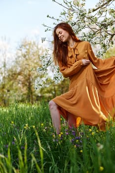 a happy, slender, sweet woman stands in a long orange dress in the tall grass near a flowering tree and happily smiling lifts the hem of her dress. High quality photo