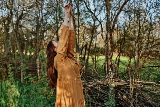 a beautiful, slender woman with long hair walks in the shade near the trees, dressed in a long orange dress, enjoying the weather and the weekend, joyfully raising her hands up. High quality photo