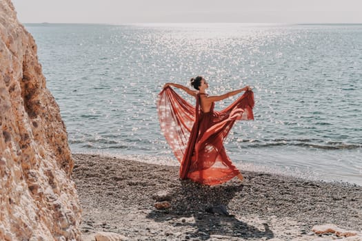 Woman red dress sea. Female dancer in a long red dress posing on a beach with rocks on sunny day. Girl on the nature on blue sky background