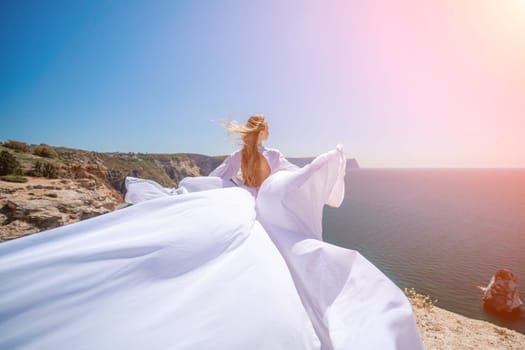 woman sea white dress. Blonde with long hair on a sunny seashore in a white flowing dress, rear view, silk fabric waving in the wind. Against the backdrop of the blue sky and mountains on the seashore