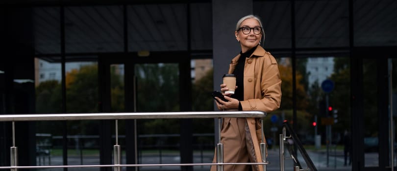 horizontal photo of a successful mature adult woman boss with a mug of coffee in her hands on the background of the building.