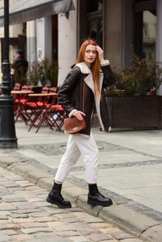 woman in white pants and leather jacket. poses outside with a small leather handbag.