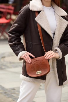 woman in white pants and a beige sweater poses outside with a small leather handbag.