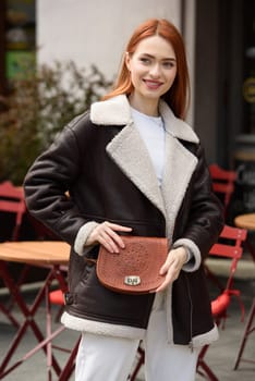 woman in white pants and leather jacket. poses outside with a small leather handbag.