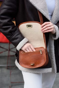 woman in white pants and a beige sweater poses outside with a small leather handbag.