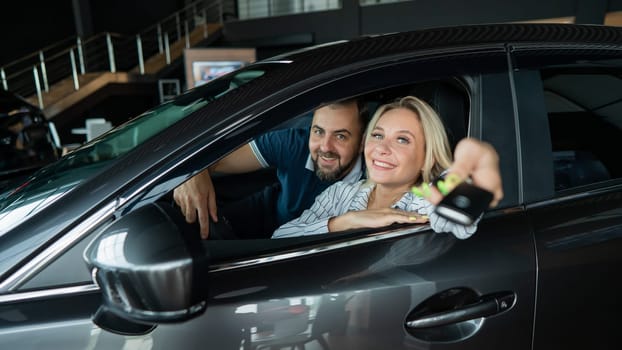 Happy caucasian couple is sitting in a car in a car dealership. A woman holds the keys to a new car