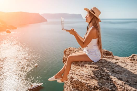 Successful business woman in yellow hat working on laptop by the sea. Pretty lady typing on computer at summer day outdoors. Freelance, travel and holidays concept.