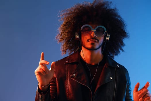 Portrait of a stylish man with curly hair with glasses and headphones on a blue background multinational, colored light, black leather jacket trend, modern concept. High quality photo