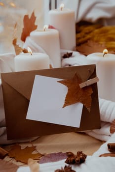 Empty paper note envelope copy space for your text on background of aesthetic atmosphere Autumn leaves spices and candle on knitted white sweater in warm yellow lights. Still life. Raining Outside