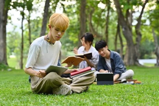 Teenage student man using digital tablet on green grass with friends sitting on background. Education, technology and lifestyle.