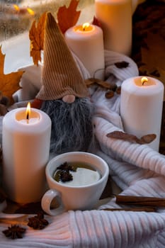 Thanksgiving and Hello Fall Halloween concept Celebrating autumn holidays at cozy home on the windowsill Hygge aesthetic atmosphere Autumn leaves gnome, spices and candle on knitted white sweater in warm yellow lights. Still life Cup of hot tea drink. Raining Outside