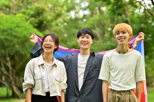 Image of young people with LGBTQ pride flag, standing in public park. LGBTQ community, freedom, solidarity and equal rights.