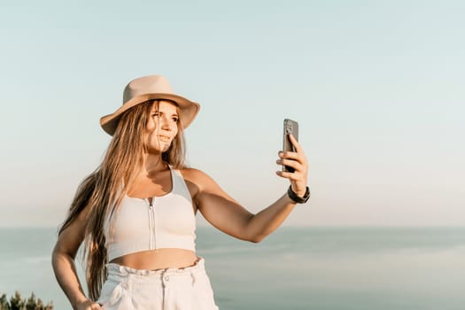 Selfie woman in hat, white tank top and shorts makes selfie shot mobile phone post photo social network outdoors on sea background beach people vacation lifestyle travel concept