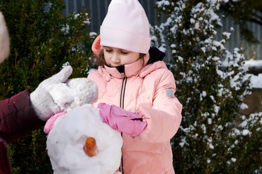 Adorable Caucasian little child girl in warm pink down jacket, having fun, enjoying making a snowman in the snow covered backyard, spending happy winter holidays outdoors. People. Leisure activity