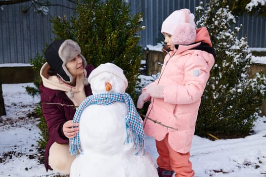 Happy mother and her adorable daughter, little child girl in pink down jacket, spending wonderful time together, building snowman in snow covered backyard. Winter leisures. Christmas holidays. Family
