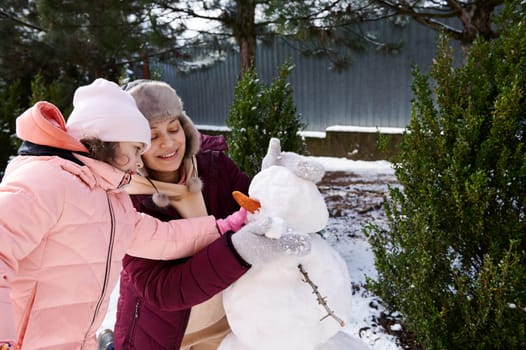 Cute little child girl playing and actively spending time on fresh frosty air with her loving mother, making snowman in a winter park. Winter holidays. Family pastime. Happy childhood. Motherhood