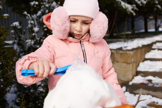 Selective focus on lovely little girl 5 years old, dressed in pink down jacket and stylish fluffy earmuffs holding a shovel and building a snowman outdoors. Leisure activities and games at wintertime