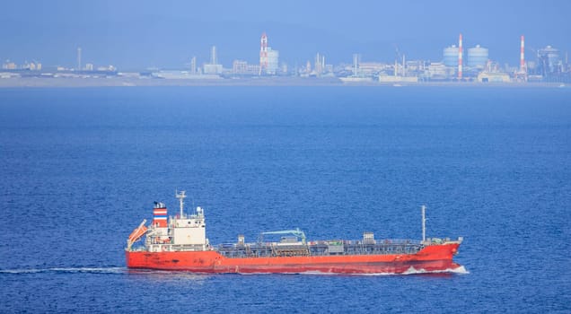 Small red cargo ship sails past chimneys at industrial refinery on coast. High quality photo