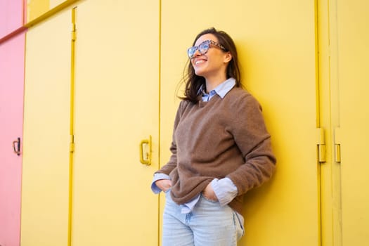 Happy woman in glasses outdoor on yellow color background side view. Positive people concept. Smiling girl looking up, hands in pocket, dressed sweater and jeans