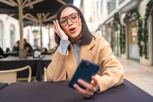 Excited middle-aged woman using smartphone sitting outdoor cafe. Surprised businesswoman in glasses dressed trench coat holding smartphone in hand looking screen