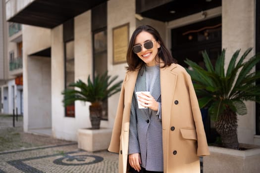 Happy caucasian girl walking with cup of coffee in city on sunny day. Brunette woman wears stylish jacket coat and sunglasses come out from building entrance. Positive emotions lifestyle concept