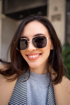 Streetstyle, street fashion concept: woman wearing trendy outfit walking in city. Cream trench coat, sunglasses. Looking camera, vertical photo close up