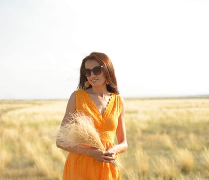 A beautiful brunette girl in an orange dress and sunglasses with a bouquet of field grass enjoys nature in the summer