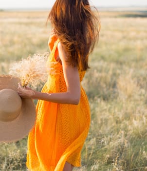 Beauty romantic girl outdoors. Rear view of a beautiful girl dressed in a casual orange dress with a straw hat in her hands on a field in the sunlight. Blows long hair. Autumn. Shine the sun, sunshine. backlit Warm tinted.