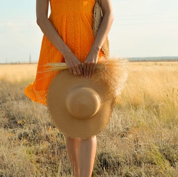 Close-up of a woman in an orange dress with a straw hat and a bouquet of field grass in her hands in a field at sunset