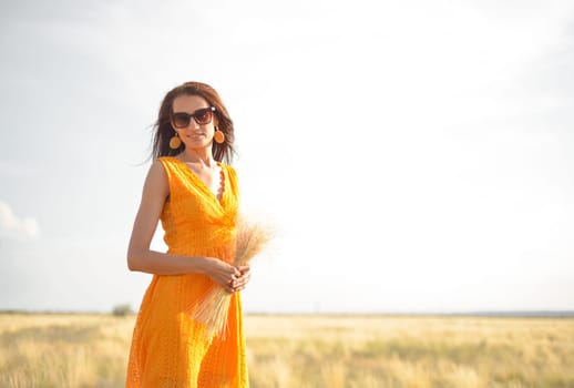 A beautiful brunette girl in an orange dress and sunglasses with a bouquet of field grass enjoys nature in the summer