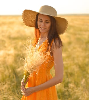 Young woman in an orange dress and a straw hat standing on a field in the rays of the setting sun.