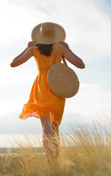 Beauty romantic girl outdoors. Rear view of a beautiful girl dressed in a casual orange dress with a straw hat and a straw bag in her hands in a field in the sunlight. Blows long hair. Autumn. Shine the sun, sunshine. backlit Warm tinted.