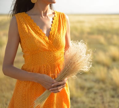 Close-up of a woman in an orange dress with a bouquet of field grass in a field at sunset