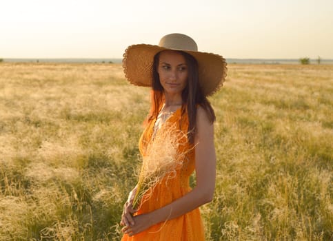 Young woman in an orange dress and a straw hat standing on a field in the rays of the setting sun.