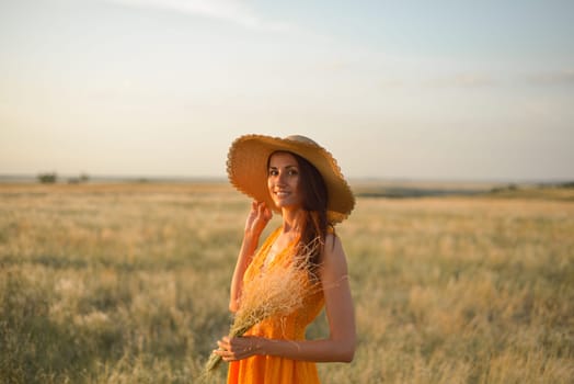 Young woman in an orange dress and a straw hat standing on a field in the rays of the setting sun.