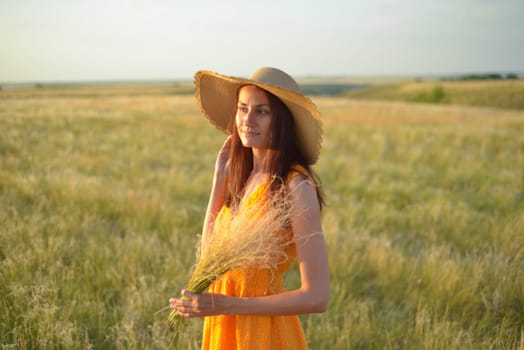 Young woman in an orange dress and a straw hat standing on a field in the rays of the setting sun.