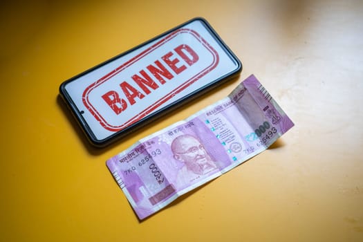 Rs 2000 Indian bank note that has been removed from circulation through demonitization on yellow table with mobile showing banned callout in India