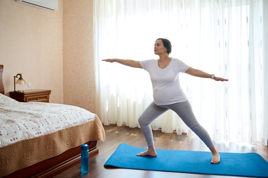 Full length portrait of multi ethnic adult pregnant woman, gravid expectant mother in late pregnancy, outstretching hands asides while practicing yoga at home, standing in warrior pose on exercise mat