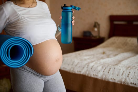 Close-up bare belly with stretch marks of a pregnant woman in gray leggins and white crop t-shirt, carrying a blue exercise mat and bottle with water. Pregnancy yoga and fitness. Childbirth. Maternity