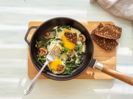 Fried eggs with spinach, mushrooms, cheese and bread in cooking pan . Healthy homemade dish for low carb diet on a light blue wooden table with linen towel. flat lay. Top view. Idea for breakfast.